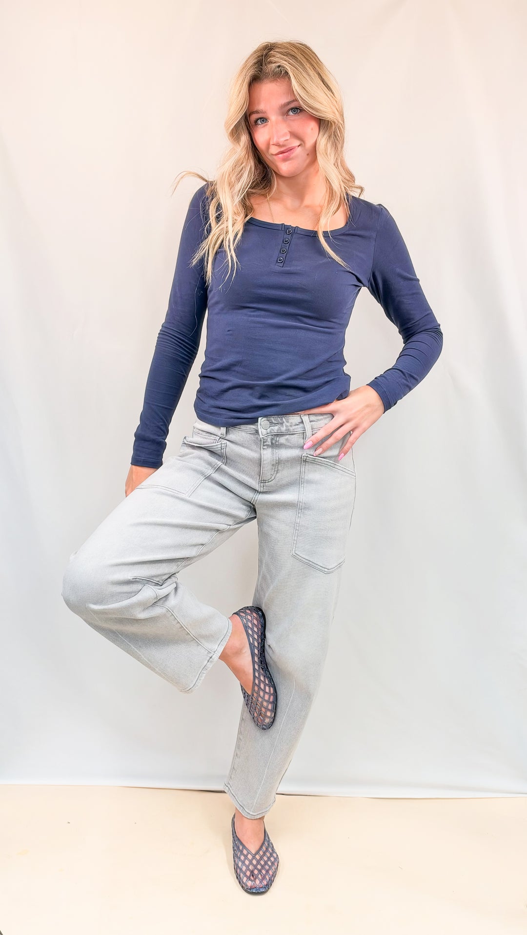 Woman wearing navy top and grey wash denim pants with blue jelly flats on plain background