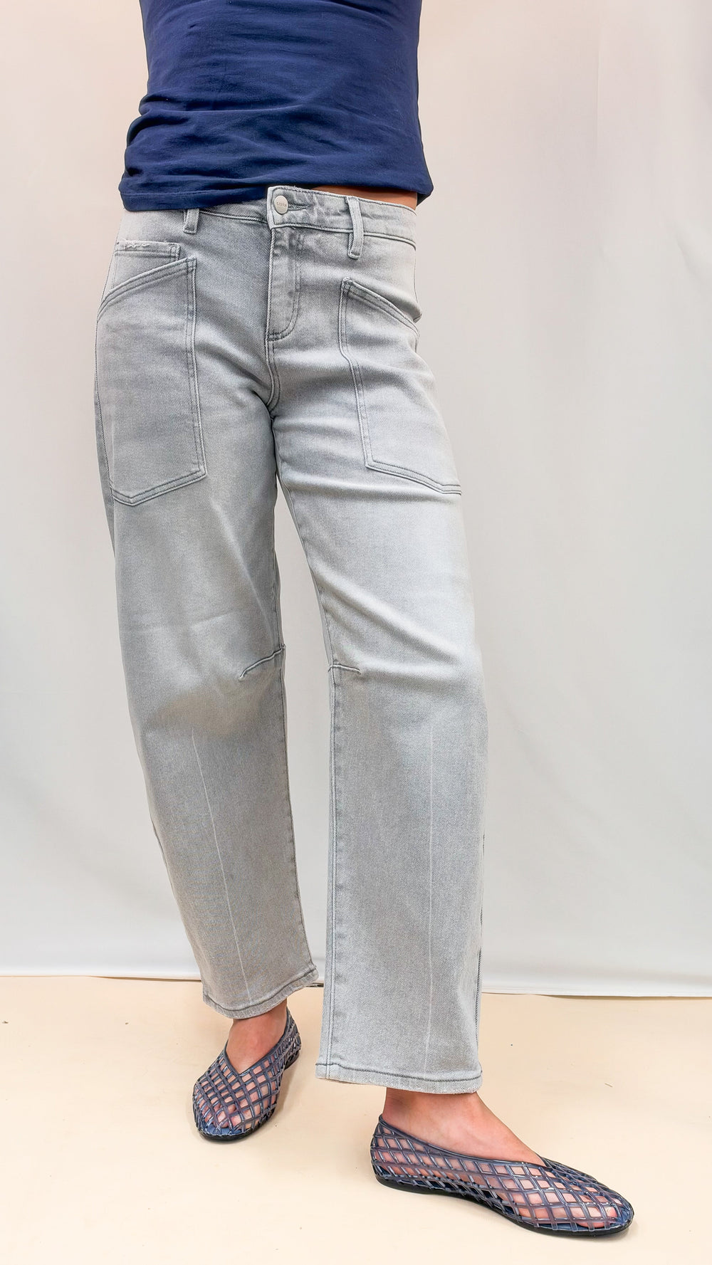 Woman wearing navy top with grey wash denim pants and blue jelly flats on plain background
