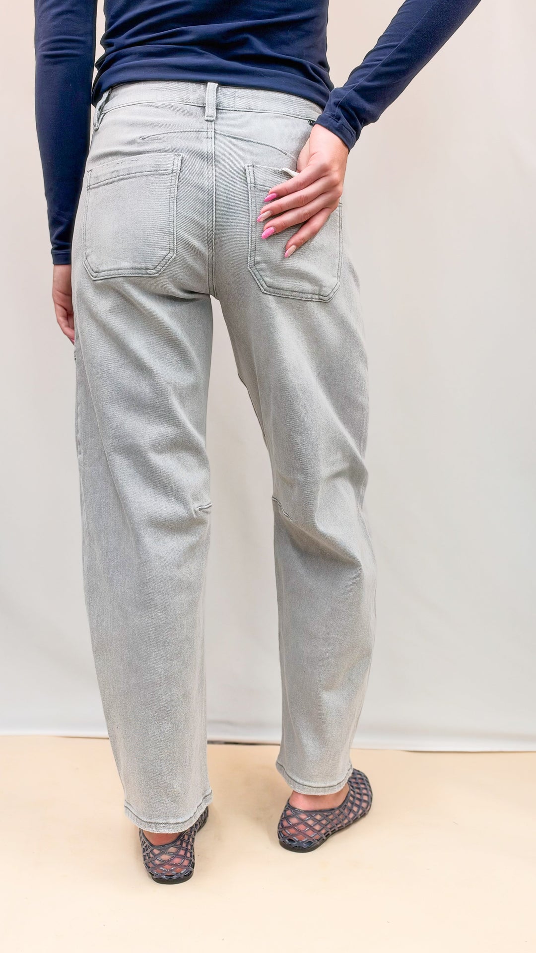 Woman shown from back wearing navy top and grey wash denim jeans with blue jelly flats on plain background