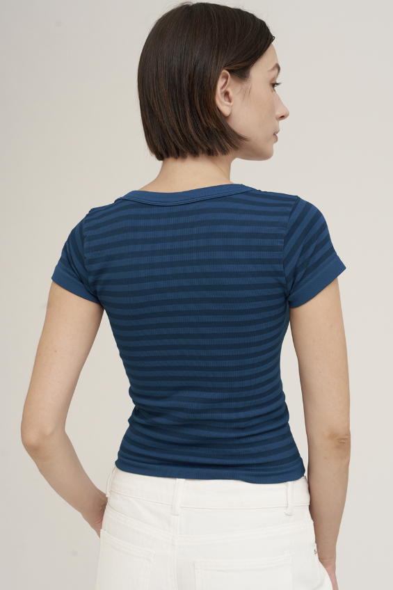 A woman shown from the back wearing a navy blue short-sleeve t-shirt with thin horizontal stripes and white pants, set against a plain light gray studio background.