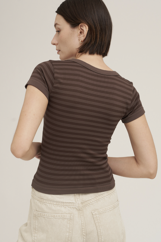 A woman shown from the back wearing a fitted short-sleeve ribbed top with horizontal dark brown and medium brown stripes, paired with light beige pants, against a plain light beige studio background.