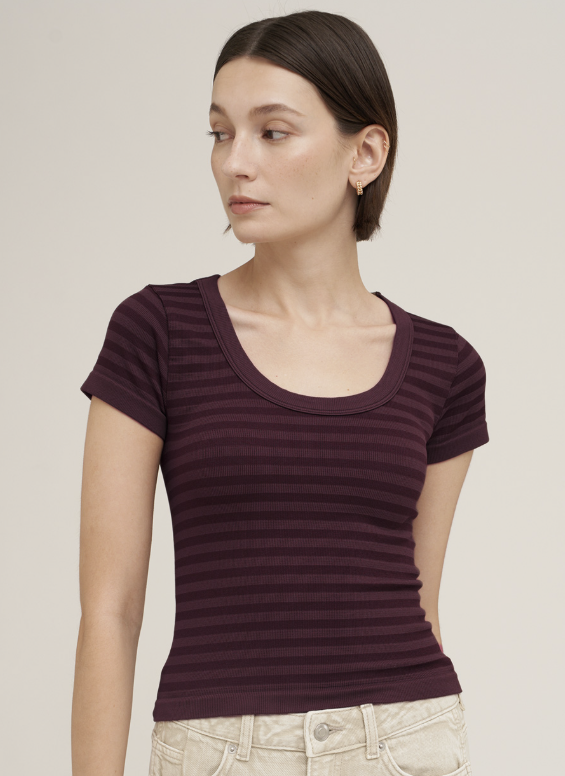 A woman wearing a short-sleeve scoop-neck ribbed top in deep plum purple, paired with light beige pants, against a solid light beige background.