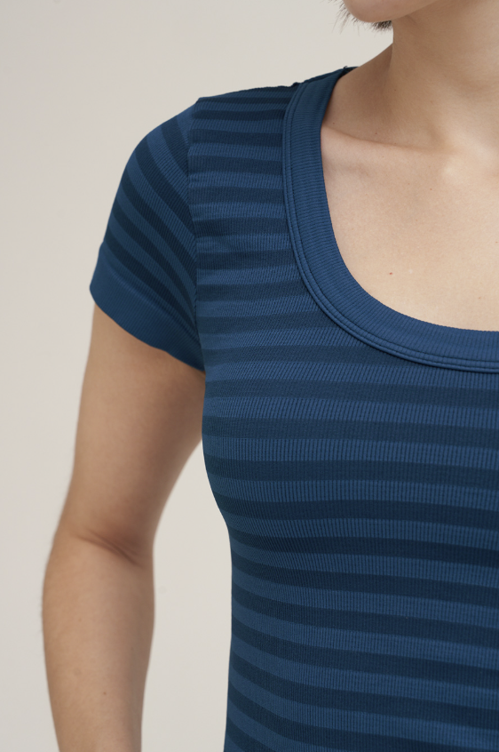 Close-up of a person wearing a short-sleeve scoop-neck top with horizontal navy and dark blue stripes against a plain light beige background.