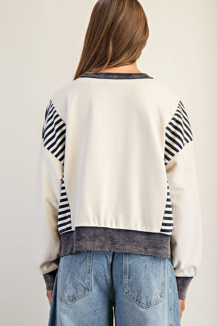 Person wearing a white sweater with black and gray striped sleeves and blue jeans on a plain background
