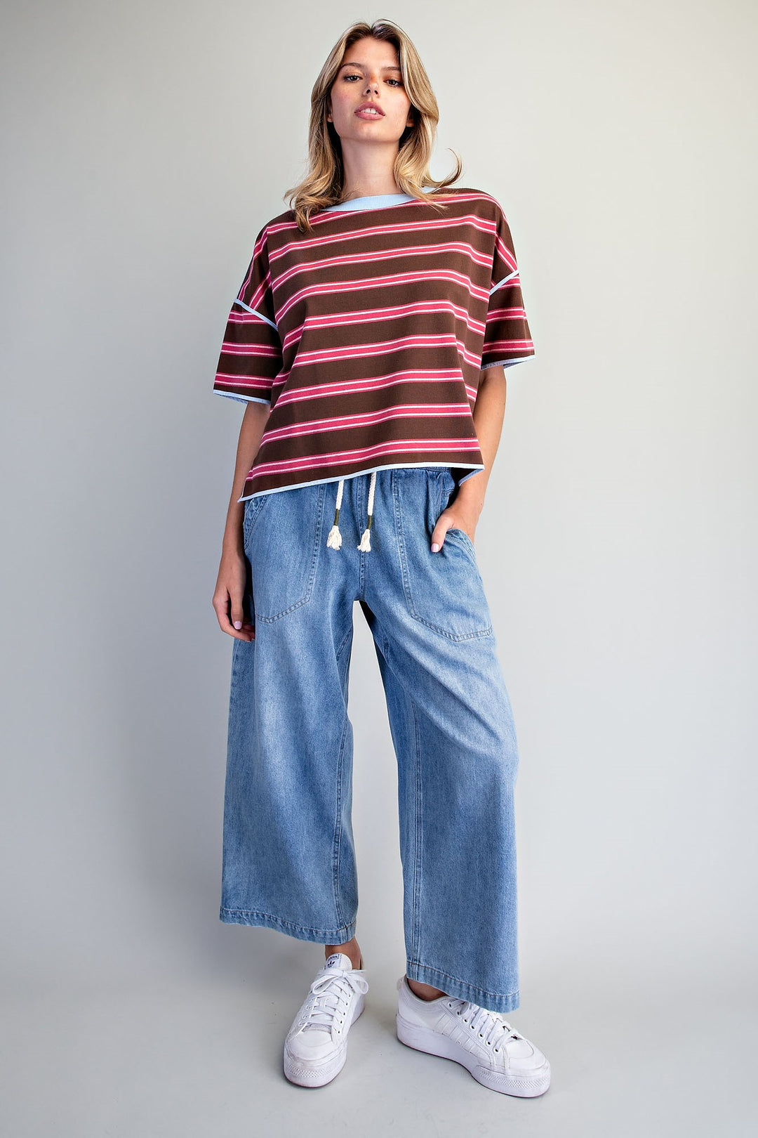 A woman modeling a short-sleeve striped t-shirt in dark brown, red, and pink, paired with wide-leg blue jeans and white sneakers, against a light gray studio background.