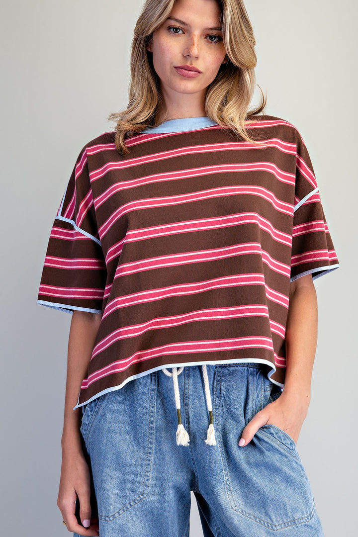 A woman wearing a brown short-sleeve crop top with pink and white horizontal stripes, paired with light blue denim drawstring pants, against a plain light gray studio background.