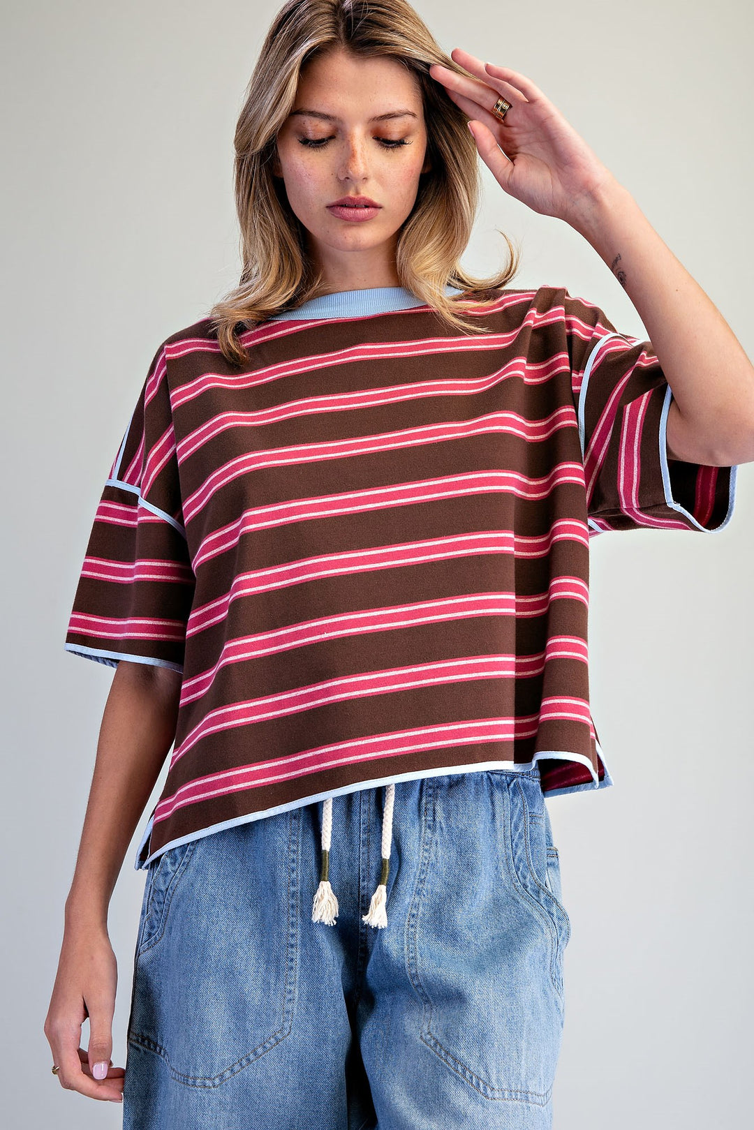 A woman wearing a brown short-sleeve crop top with pink and white horizontal stripes, paired with light blue denim jeans, against a plain light gray background.