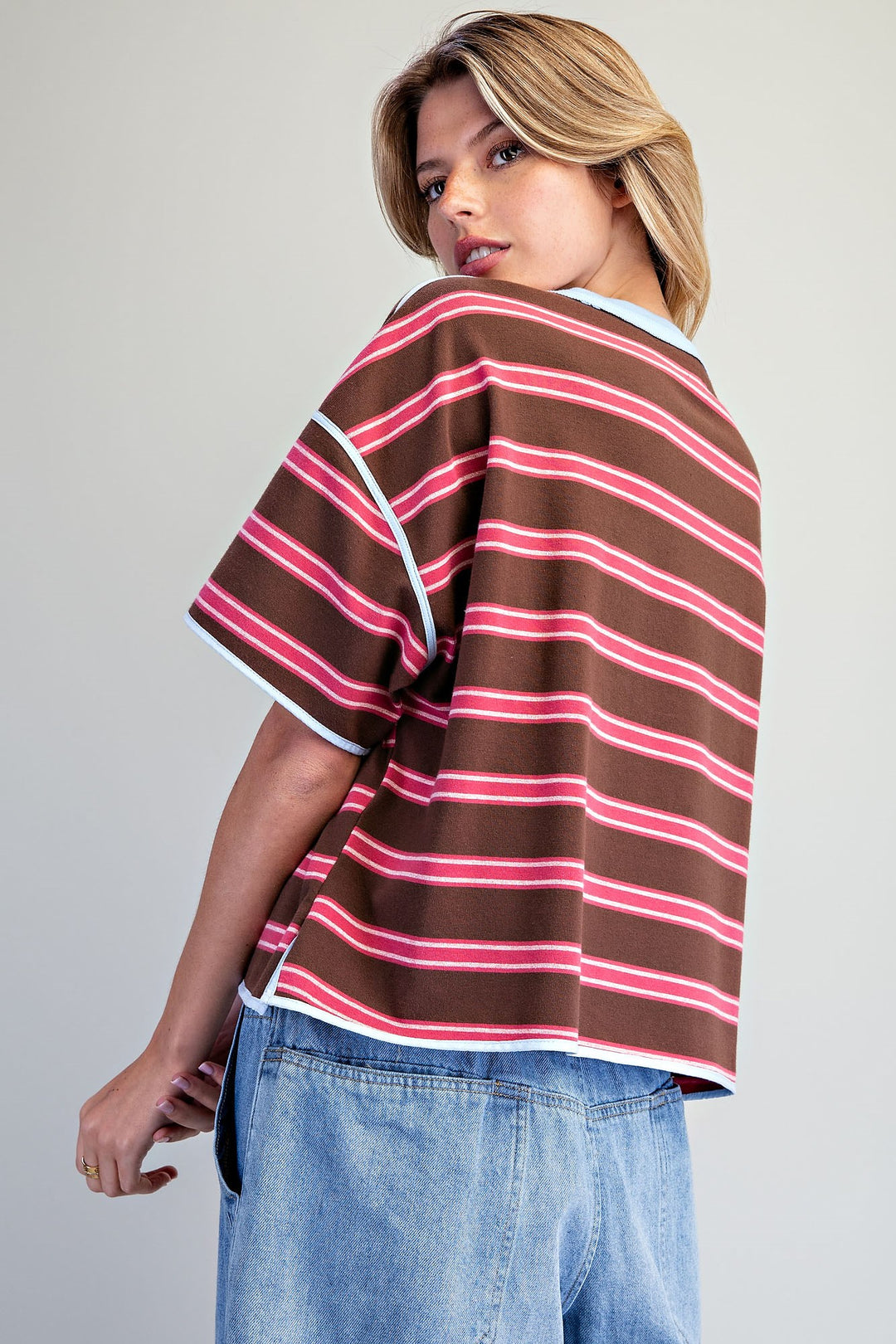 A woman wearing a brown short-sleeve striped knit top with pink and white stripes, paired with light blue jeans, photographed against a plain light gray studio background.