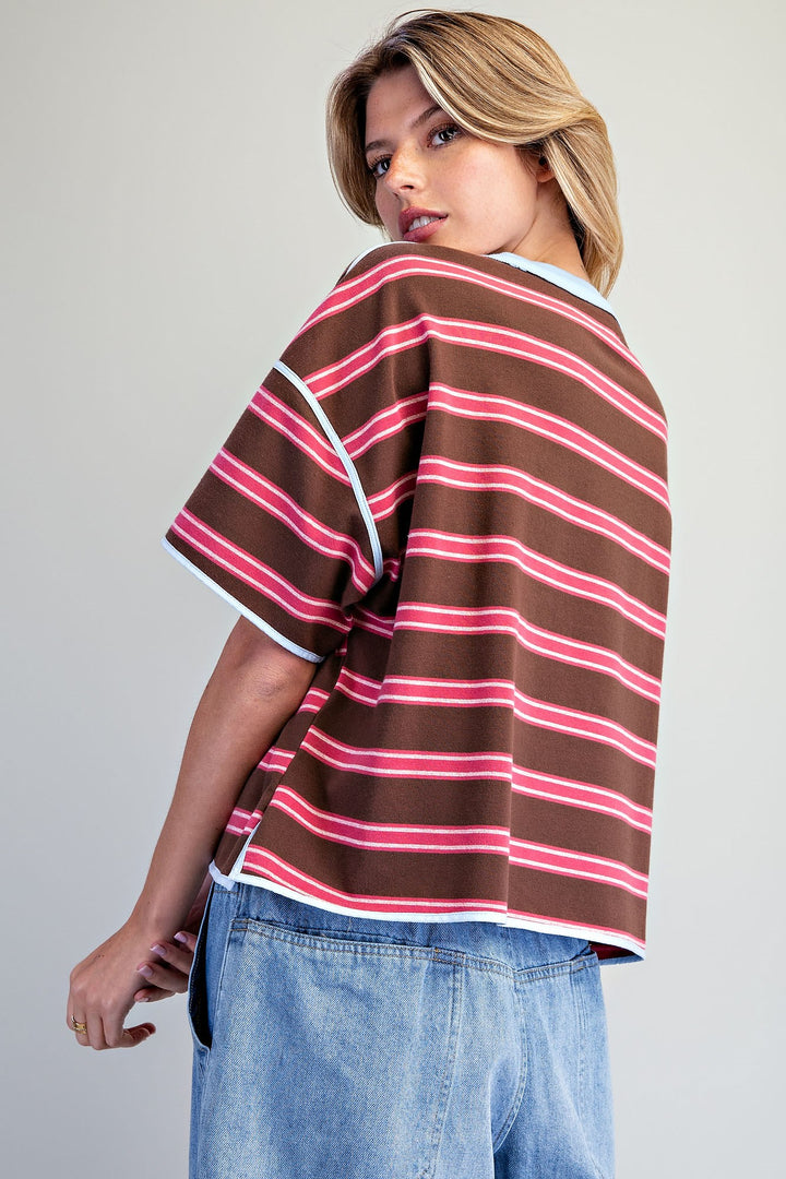 A woman wearing a brown short-sleeve striped knit top with pink and white stripes, paired with light blue jeans, photographed against a plain light gray studio background.