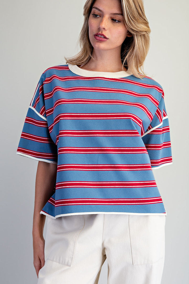 A woman wearing a blue short-sleeve striped top with red and white horizontal stripes, paired with white pants, against a plain light gray background.