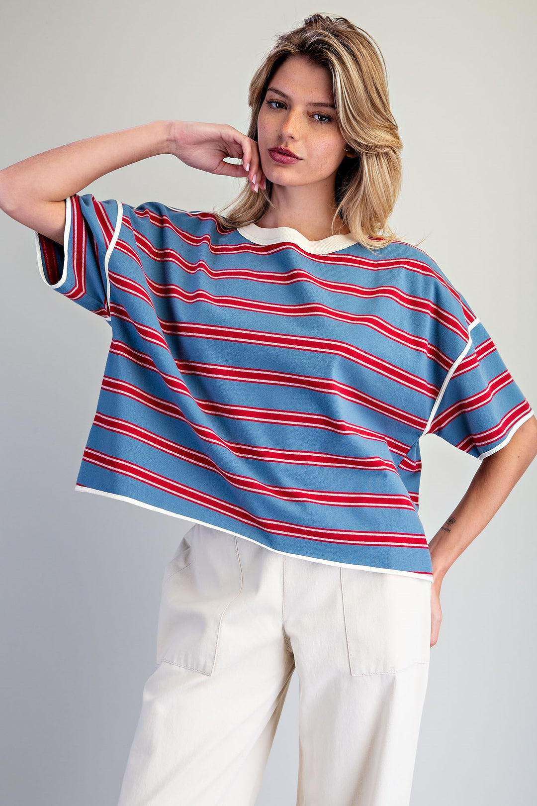 A woman modeling a cropped short-sleeve t-shirt with horizontal blue, red, and white stripes, paired with off-white trousers, against a plain light gray studio background.