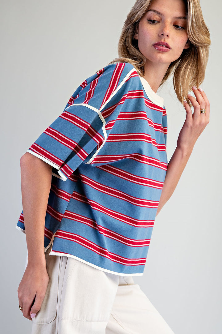 A woman wearing a blue short-sleeve striped top with red and white horizontal stripes, paired with off-white pants, photographed against a light gray studio background.