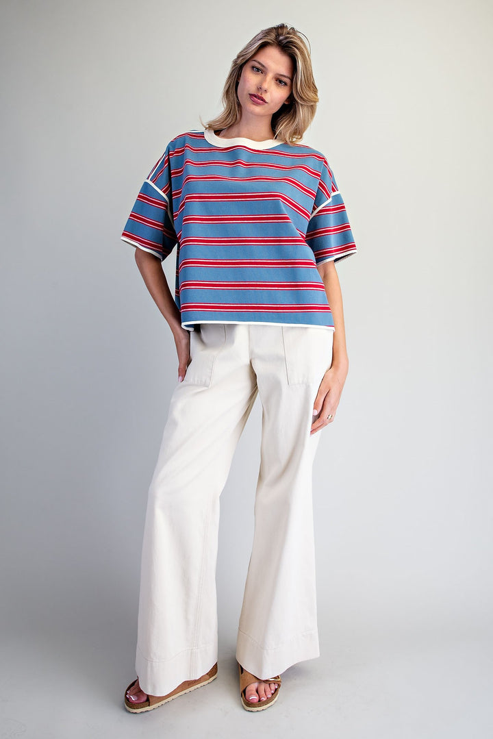 A woman modeling a short-sleeve striped knit top with red, blue, and white horizontal stripes, paired with off-white wide-leg pants and brown sandals, against a light gray studio background.