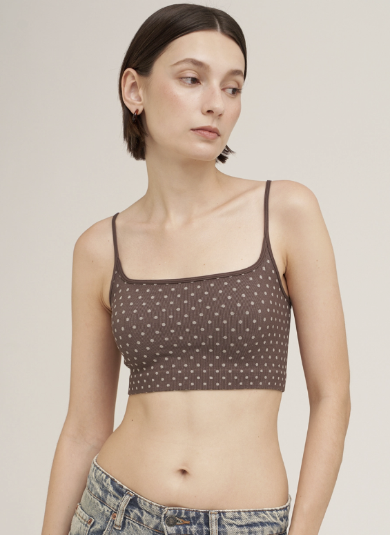 A woman wearing a brown crop top with a small white polka dot pattern and thin straps, paired with blue jeans, against a solid light beige background.