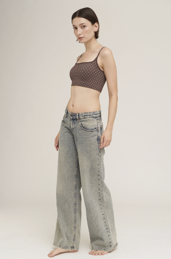 A woman modeling a brown and white polka dot crop top with thin straps and light-wash wide-leg denim jeans against a plain light beige studio background.