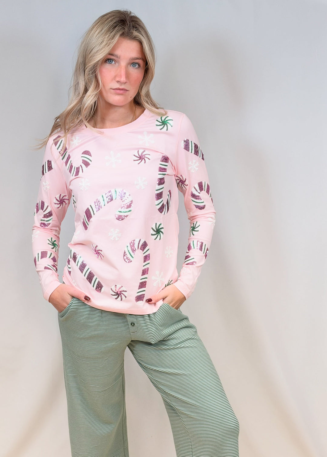 Woman wearing pink long sleeve top with sequin candy canes and candies with green stripe pants on plain background