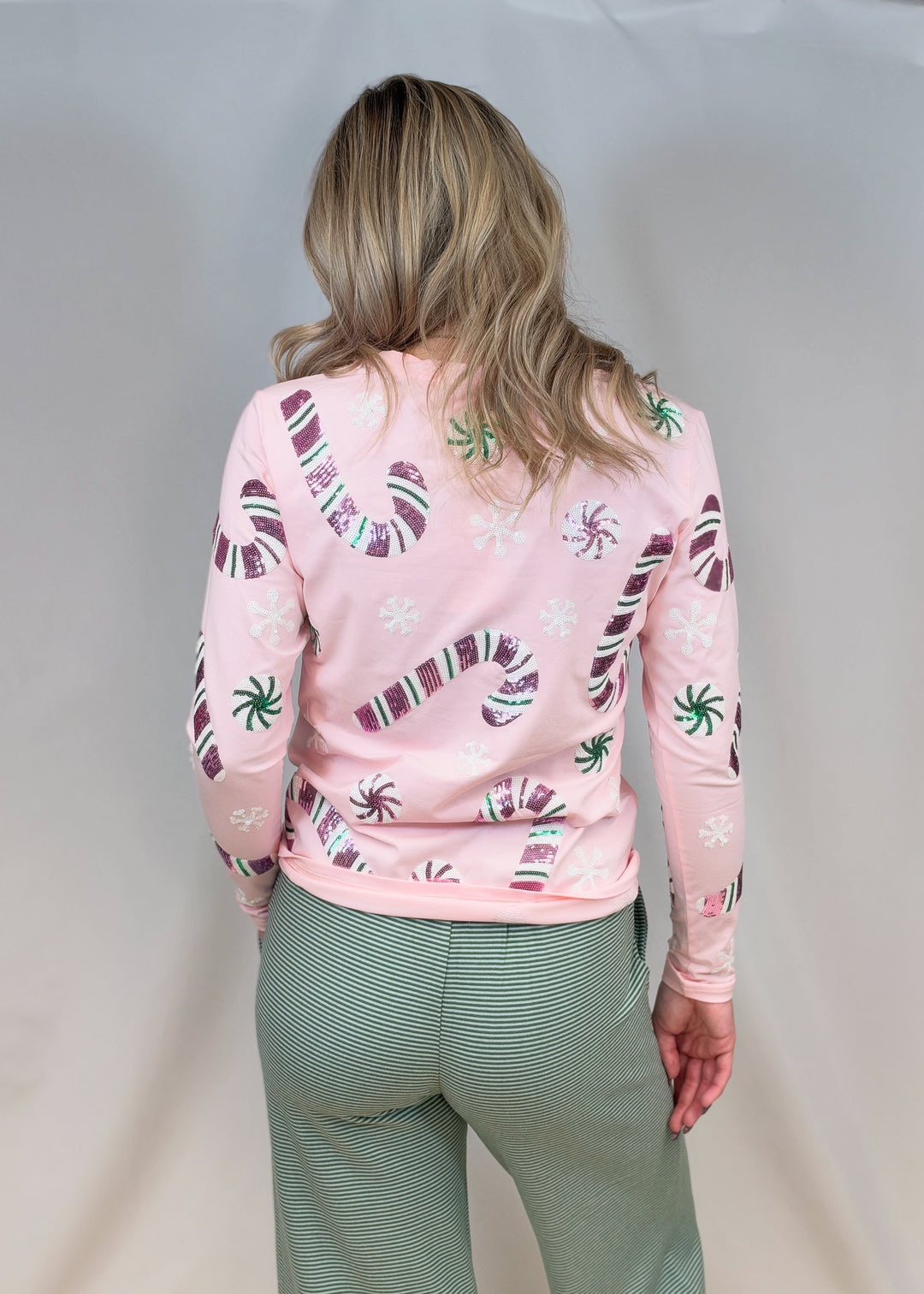 Woman shown from back view wearing pink long sleeve top with sequin candy canes and candies with green stripe pants on plain background