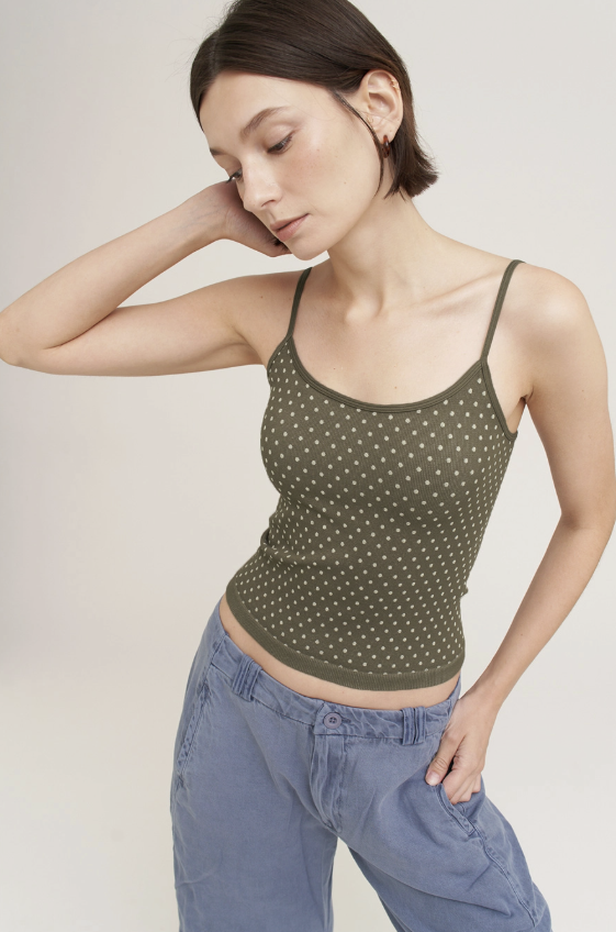 A woman wearing a fitted olive green spaghetti-strap tank top with small white polka dots, paired with light blue jeans, photographed against a plain light beige studio background.
