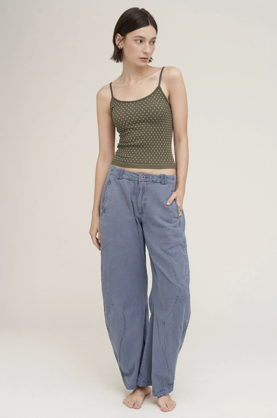 A model stands barefoot against a plain light beige studio background, wearing a sleeveless olive green top with a small repeating pattern and loose blue wide-leg pants.