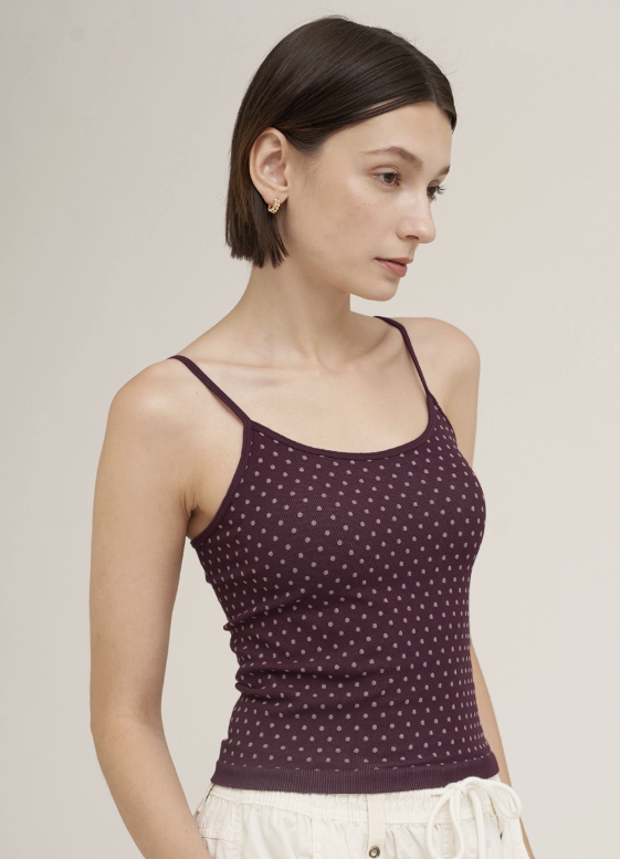 A woman wearing a dark plum purple ribbed knit camisole top with a small white polka dot pattern, paired with white drawstring pants, photographed against a solid light beige background.
