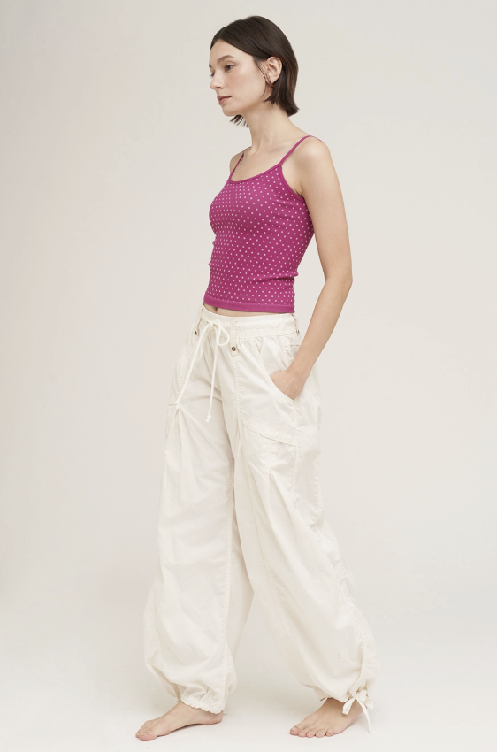A woman stands barefoot against a plain light beige studio background, wearing a sleeveless pink top with a small repeating pattern and loose white drawstring pants.