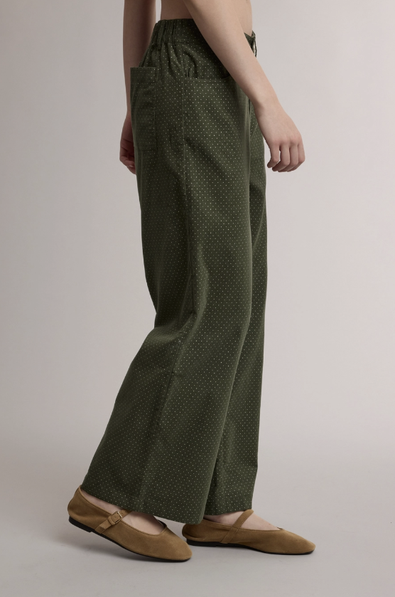 A model is shown from the waist down wearing olive green wide-leg trousers with a small white polka dot pattern and tan ballet flats, set against a plain light gray studio background.