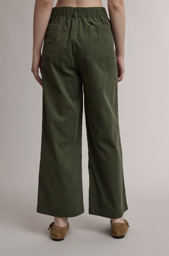 A person shown from the back wearing olive green wide-leg pants with a small white geometric pattern, paired with tan suede mules, against a neutral studio background.