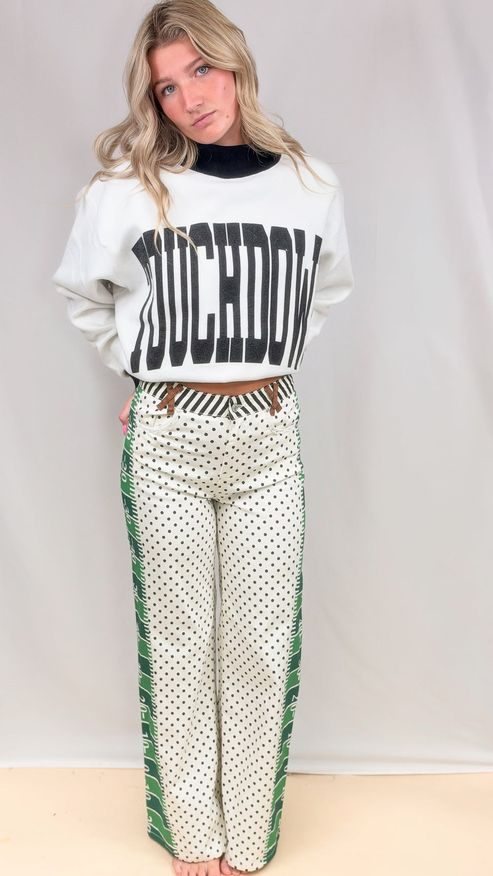 Woman wearing white and black mock neck with 'TOUCHDOWN' on front with football field pattern pants on plain background