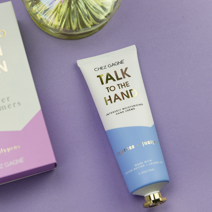 A tube of Chez Gagnée Talk to the Hand moisturizing hand cream, featuring a white and light blue design with gold lettering, placed on a lavender background next to its packaging box.