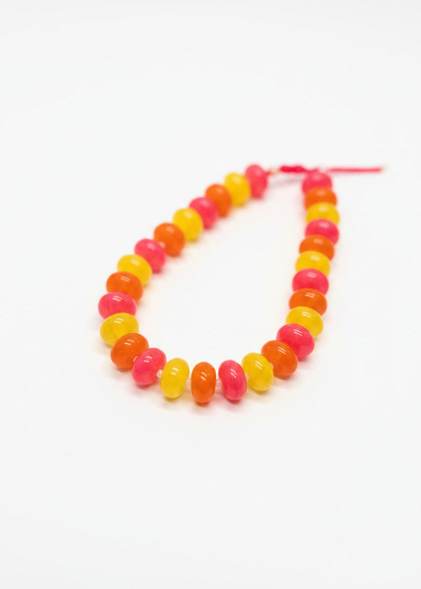 A beaded necklace made of alternating translucent pink, orange, and yellow beads, displayed on a white card over a dark patterned fabric background.