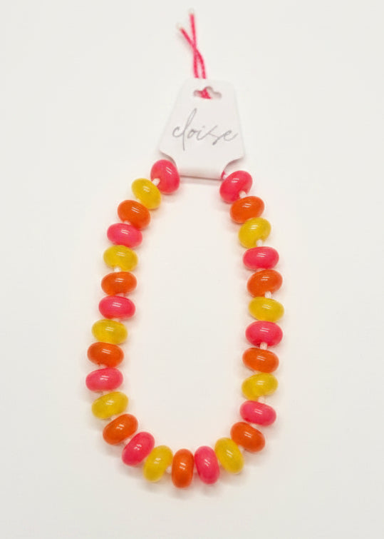 A beaded necklace made of alternating bright pink, orange, and yellow oval beads, displayed on a white card held by a hand over a green patterned carpet.