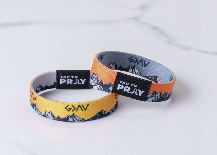 Two wristbands with a mountain pattern and 'TAP TO PRAY' labels, shown on a light marble background. One wristband is yellow, and the other is orange.