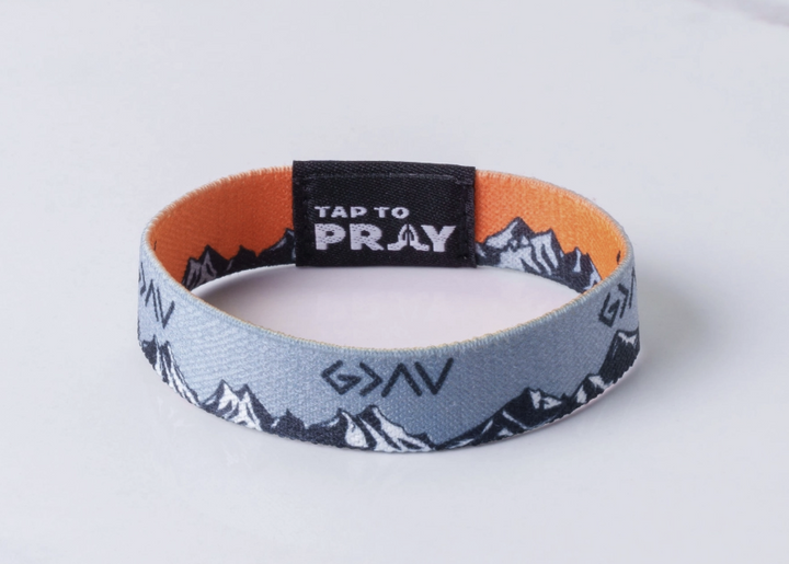 A fabric wristband bracelet with a light blue and black mountain pattern, an orange inner lining, and a black closure tab that reads &