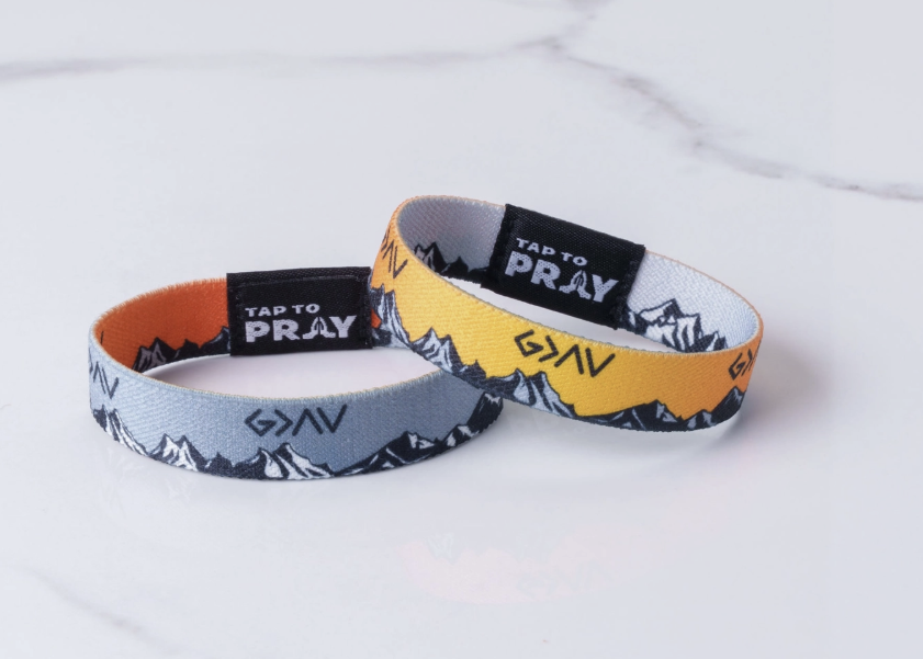 Two silicone wristbands with a mountain pattern, one in gray and one in yellow, displayed on a white marble surface. Both bands feature a black inner section with the brand name 'TAP TO PRAY' and a black logo, and the outer section features a gray and yellow mountain design.