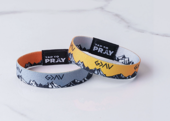 Two silicone wristbands with a mountain pattern, one in gray and one in yellow, displayed on a white marble surface. Both bands feature a black inner section with the brand name &