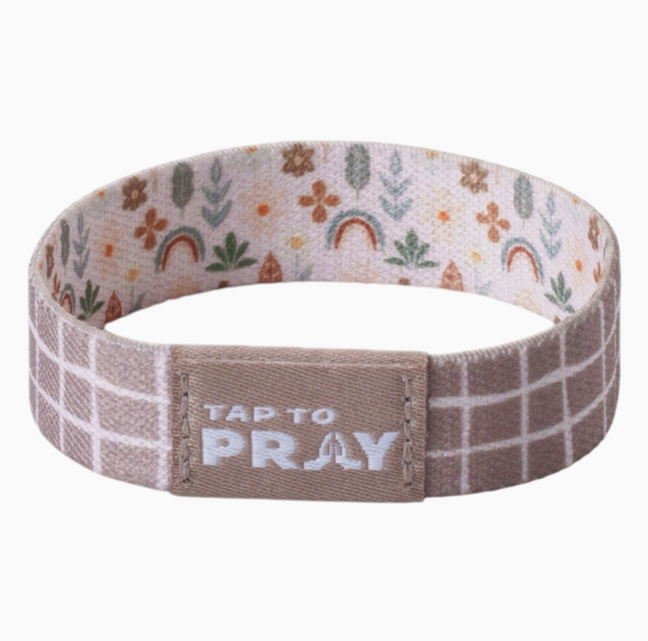 A fabric wristband with a brown and white plaid pattern on the outside, and a light pink patterned lining with small flowers, rainbows, and leaves. The center has a brown tab with the text &