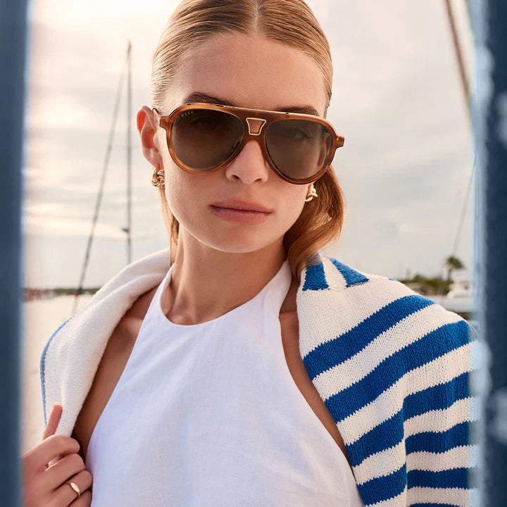 Woman wearing sunglasses and a blue and white striped cardigan by a body of water.