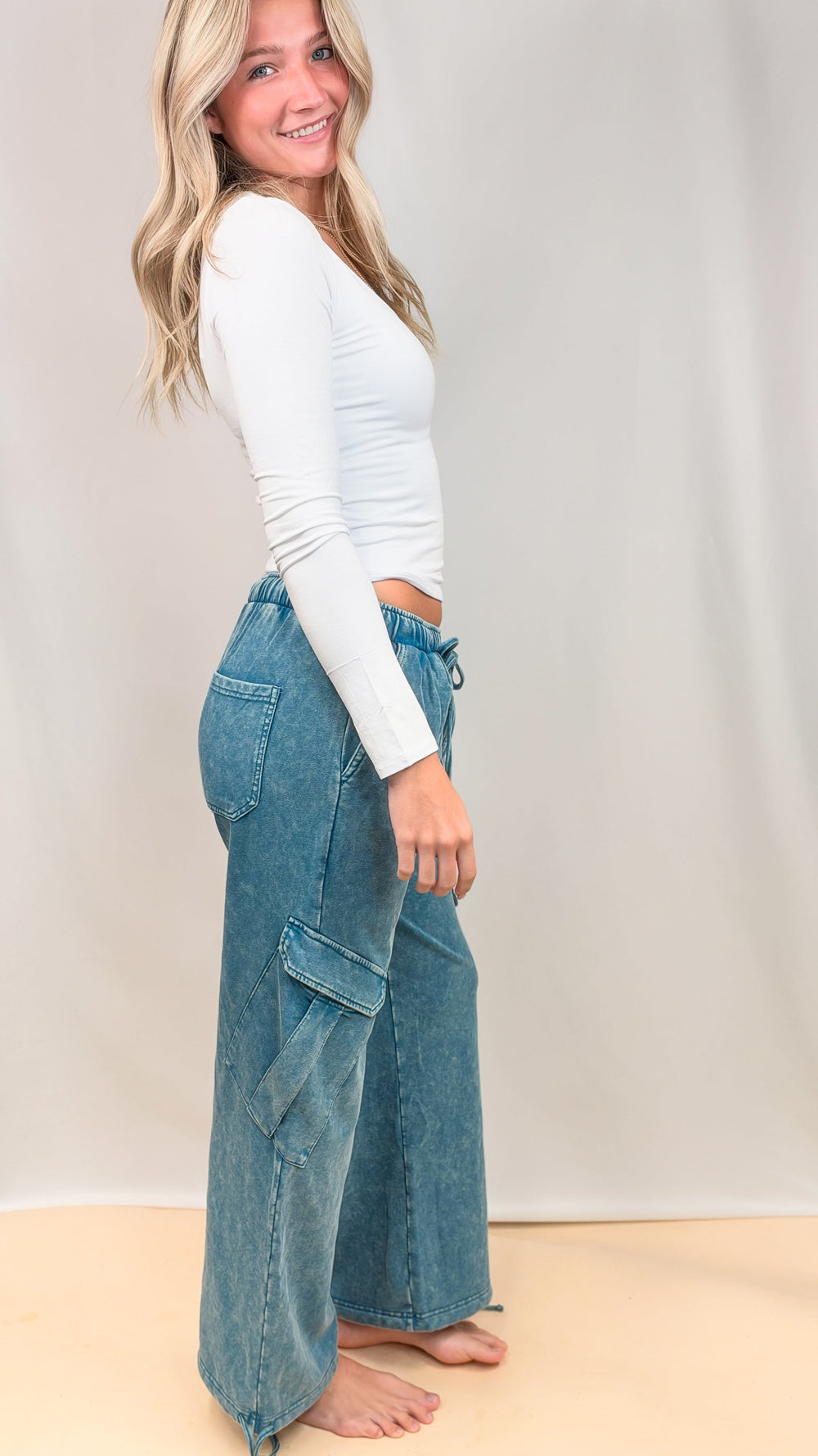 Woman shown from side view wearing white top and teal blue pants on plain background