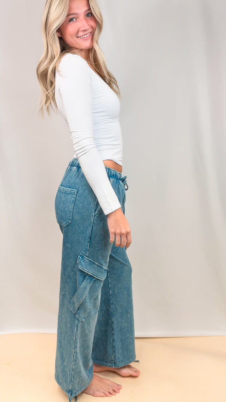 Woman shown from side view wearing white top and teal blue pants on plain background
