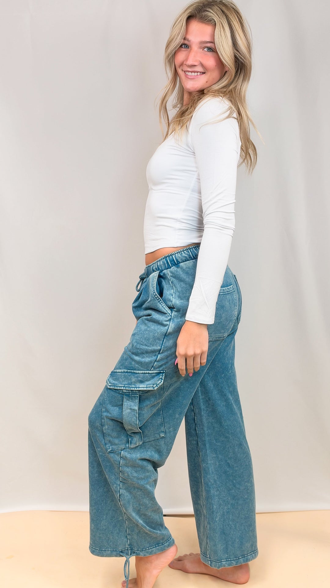 Woman shown from side view wearing white top and teal blue pants on plain background