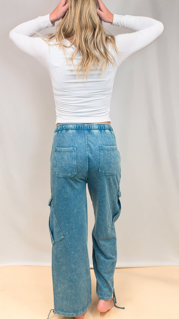 Woman shown from back view wearing white top and teal blue pants on plain background