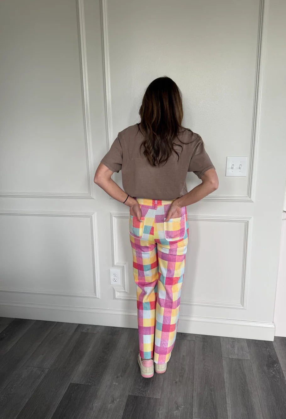 Person wearing a brown shirt and colorful checkered pants standing in a room with white paneled walls.