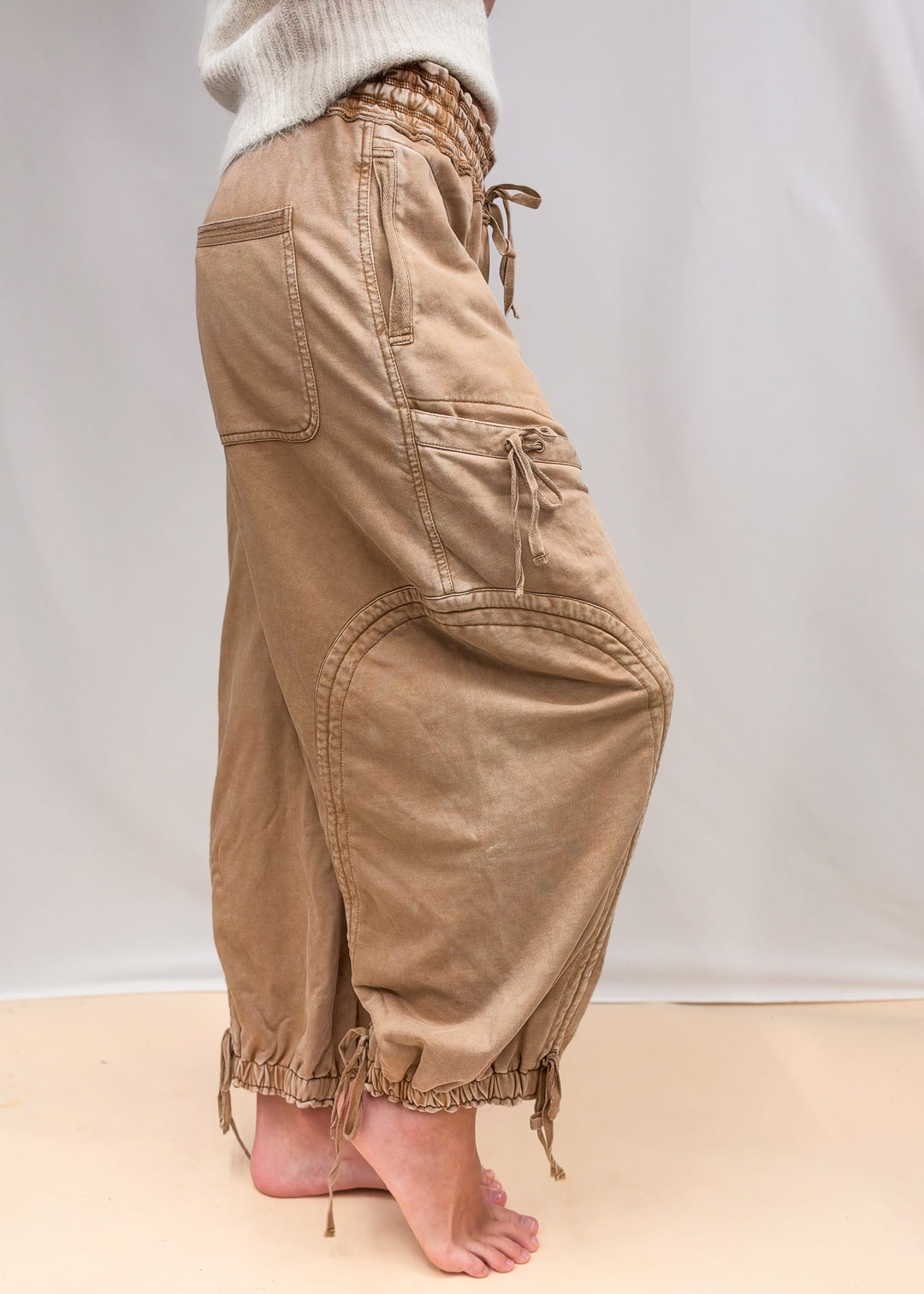 Woman wearing camel color sweatpants with drawstring details and cream top on plain background