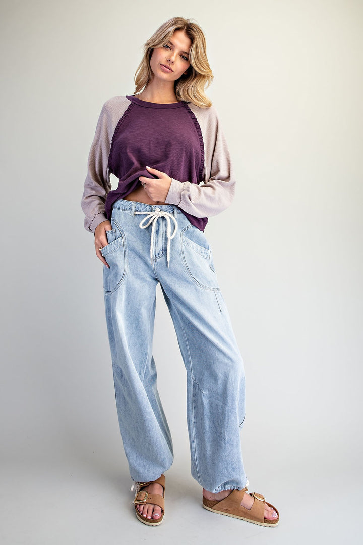 A woman modeling a two-tone raglan long-sleeve top with a deep purple crew neck and light beige sleeves, paired with wide-leg light blue jeans and brown slide sandals, against a plain light gray studio background.