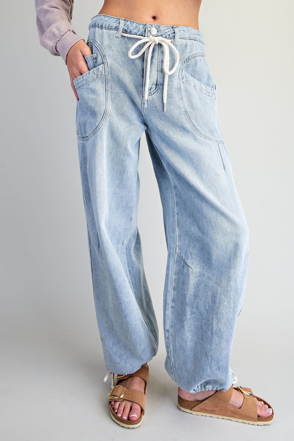 A person wearing light-wash wide-leg denim jeans with a white drawstring waist, shown from the waist down against a plain light gray studio background. The person is also wearing tan slide sandals.