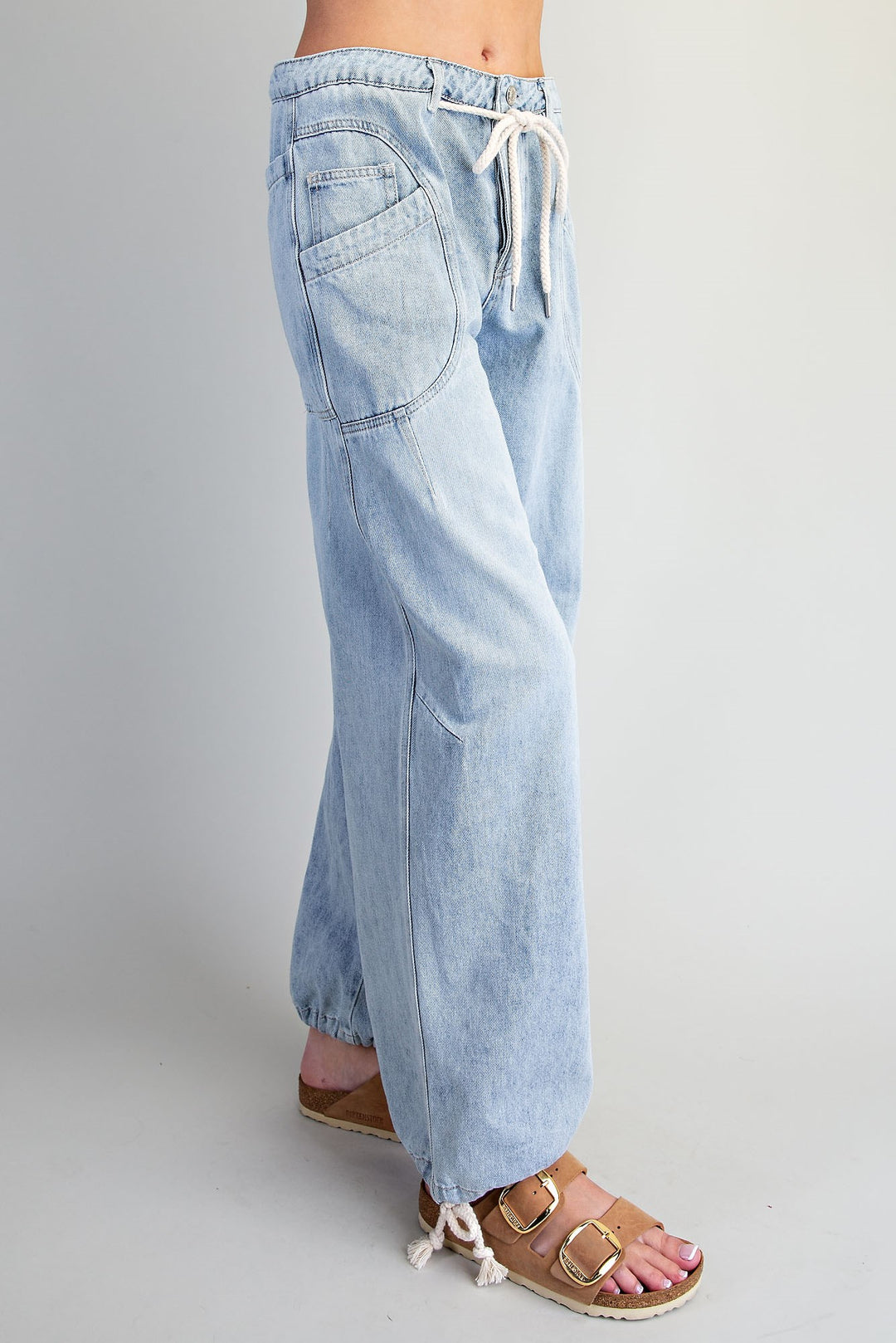 Light wash blue denim wide-leg pants with an elastic drawstring waist and side pockets, shown on a model from the waist down wearing brown sandals against a light gray studio background.