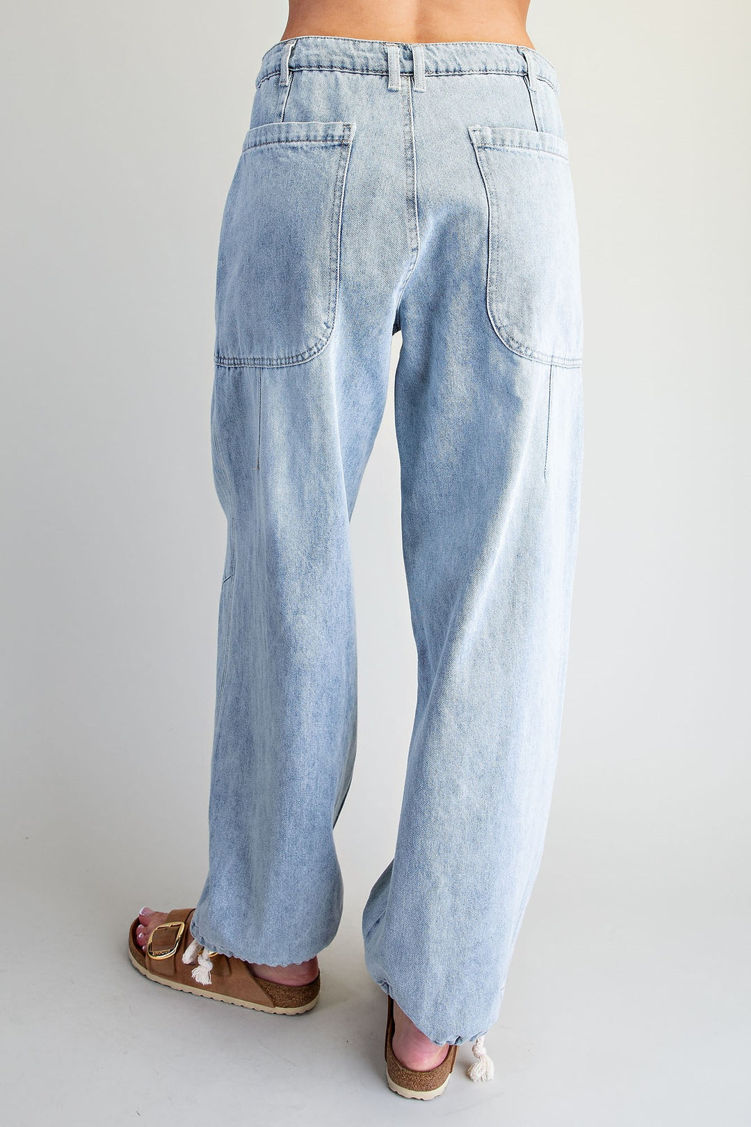 A person shown from the back wearing light-wash, wide-leg denim jeans and brown espadrille sandals against a plain light gray studio background.