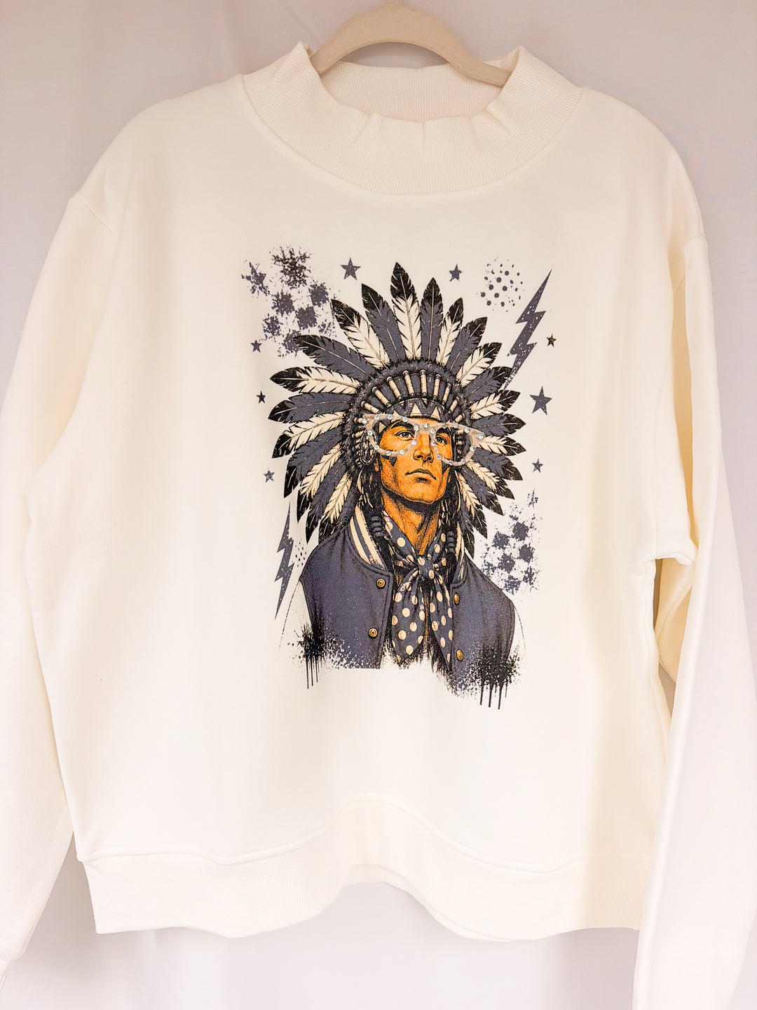 An off-white crewneck sweatshirt hanging on a wooden hanger, featuring a graphic print of a man in a Native American style headdress with stars and lightning bolts.