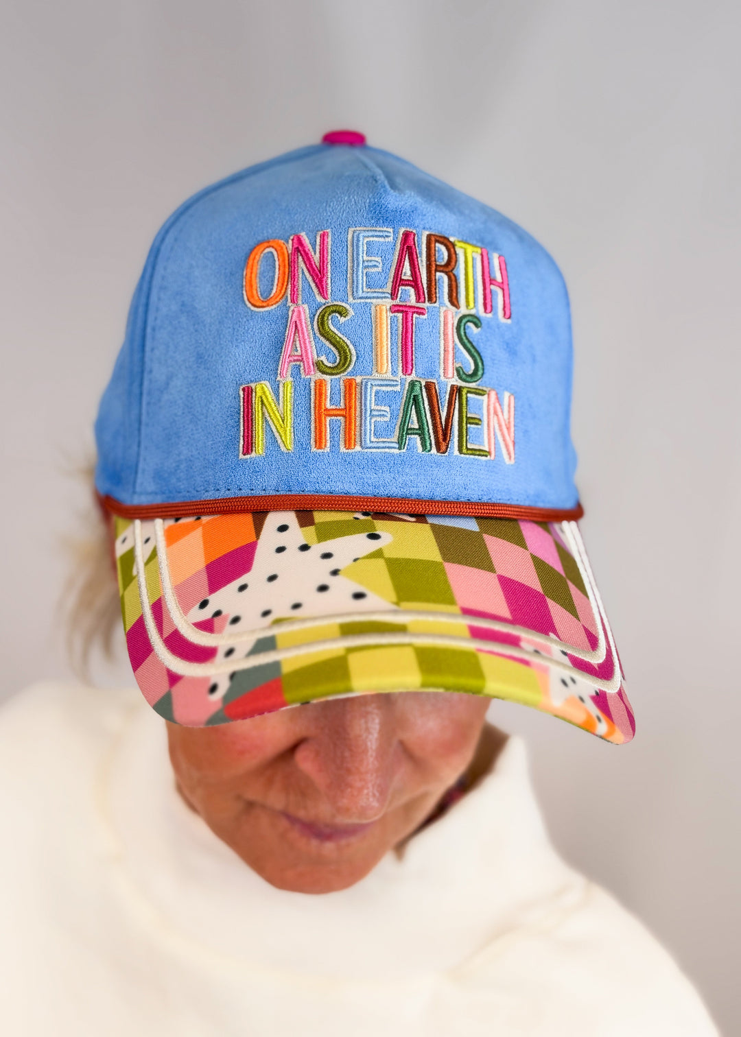 Woman wearing blue suede trucker hat with 'ON EARTH AS IT IS IN HEAVEN' embroidered on front on plain background