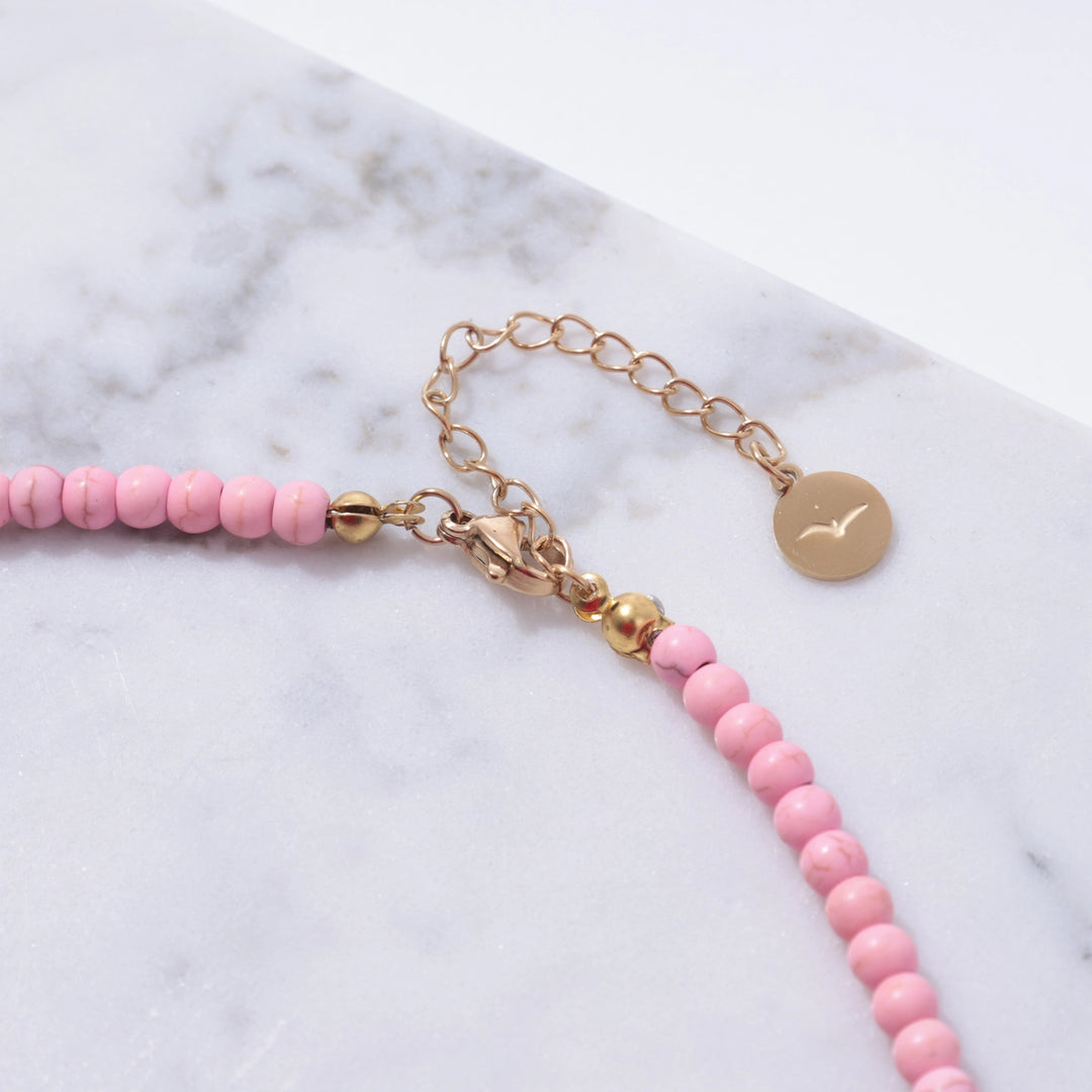 Pink beaded bracelet with gold chain and disc charm on a marble surface
